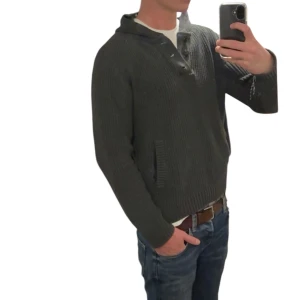 Mörkgrå stickad hoodie med knappar - Säljer en mörkgrå stickad hoodie med huva och knappar vid halsen. Tröjan har ribbade muddar och fickor på sidorna, perfekt för dig som gillar en chill och avslappnad stil. Passar dig som vill ha något mjukt och varmt med snygga detaljer.