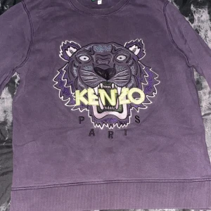 Lila Kenzo sweatshirt med tiger - Säljer en lila sweatshirt från Kenzo med ikonisk broderad tiger och logga på bröstet. Tröjan har rund hals, ribbade muddar och är långärmad. Perfekt för dig som vill sticka ut med ett coolt statement-plagg från Kenzo Paris.