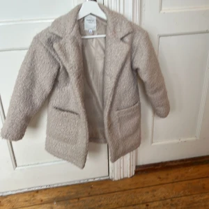 Beige teddyjacka från Name It, stl 128 - Mysig beige teddyjacka från Name It i storlek 128. Jackan har klassisk krage, två fickor framtill och är helfodrad. Perfekt för dig som vill ha en varm och trendig look med lite oversized känsla.