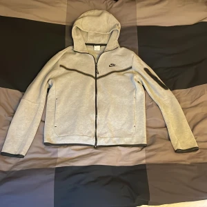 Grå hoodie från Nike med dragkedja - Snygg ljusgrå hoodie från Nike i storlek M. Tröjan har hel dragkedja framtill, huva och svarta detaljer vid dragkedjor och muddar. Nike-logga i svart på bröstet och en extra ficka med dragkedja på ena ärmen. Perfekt för chill eller träning. Skriv om intresse. Vi kan prata om priset.