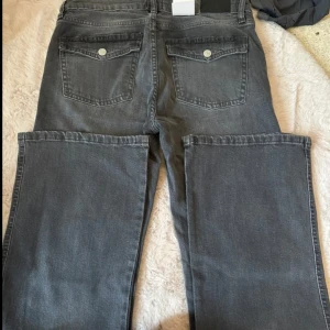 Grå bootcut jeans från 157 - Säljer ett par gråa bootcut jeans från 157 med breda ben och coola fickor med lock bak. Jeansen har klassisk femficksdesign, hällor för bälte och stängs med knapp och dragkedja. Perfekta för dig som gillar en avslappnad och trendig look.