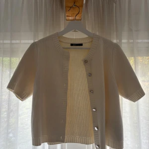 Beige stickad kofta från Gina Tricot - Kortärmad beige stickad kofta från Gina Tricot med ribbade detaljer och knappar framtill. Perfekt för lager-på-lager och har en clean, enkel look som passar till allt. Snygg att styla över linne eller t-shirt.