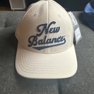 Beige keps från New Balance - Snygg och trendig keps från New Balance i stilren beige/cream nyans med mörkblå broderad logga framtill. Justerbar snapnack rem, one size. Bra passform, hög kvalitet, helt oanvänd, Tagg sitter kvar. Material: 97% Polyster, 3% spandex.