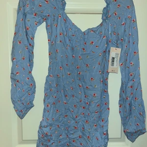 Blommig blå kortklänning från Nelly - Supersöt blå kortklänning från Nelly med små röda blommor över hela tyget. Klänningen har långärmade puffärmar, v-ringad hals och lätt rynkade detaljer. Perfekt för dig som gillar romantisk och feminin stil. Materialet är mjukt och känns skönt mot huden.