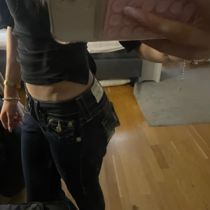 Mörkblå True Religion jeans med kontrastsömmar - Säljer ett par mörkblå jeans från True Religion med guldiga detaljer på fickorna. Byxorna har låg midja och klassisk bootcut-modell. Ikonisk broderad logga på bakfickan och coola detaljer framtill. Perfekta för dig som vill sticka ut. Omsydda till bootcut. 