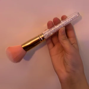Glittrig sminkborste med rosa borst - Superfin sminkborste med fluffiga rosa syntetborst, guldig metallhylsa och ett transparent handtag fyllt med glittrande kristaller. Perfekt för att applicera puder, bronzer eller rouge och ger en lyxig känsla till sminkrutinen.