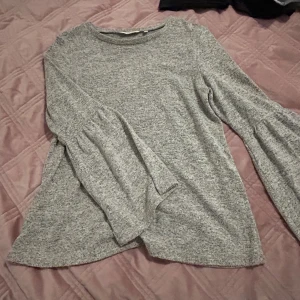 Grå långärmad tröja från Happy Holly - Supermjuk gråmelerad tröja från Happy Holly med vida, lätt utsvängda ärmar och rund halsringning. Perfekt för dig som gillar en chill och snygg look. Materialet känns mysigt och passar till många outfits.