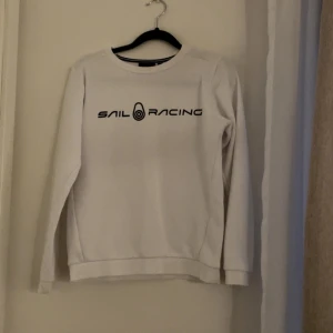 Vit Sail Racing sweatshirt - Vit sweatshirt från Sail Racing med svart logga tryckt över bröstet. Tröjan har rund halsringning med svart-vita ränder på insidan och långa ärmar. Perfekt för en clean och sportig stil.