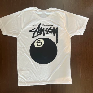 Vit Stüssy t-shirt med 8-ball print - Vit t-shirt från Stüssy med stort svart 8-ball tryck och Stüssy-logga på ryggen samt mindre print på bröstet. Klassisk passform och rund hals. 