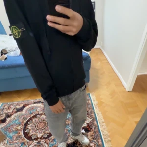 Svart hoodie från Stone Island  - Svart hoodie från Stone Island med ikonisk patch på vänster ärm. Tröjan har huva med snörning, dragkedja framtill och är tillverkad i mjuk bomull. Perfekt för dig som vill ha en clean och stilren look med street-vibe.