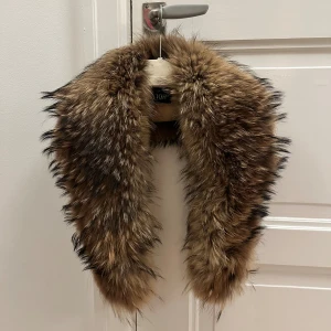 Brun pälskrage - Stor pälskrage från tvättbjörn i toppskick, ca 100cm lång. Mycket fluffig och jämn i pälsen.  Har två klämmor för att enkelt knäppa fast på t.ex jacka eller runt halsen som en halsduk.