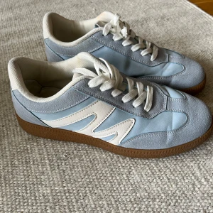 Blå och vita sneakers med mockadetaljer - Snygga sneakers i ljusblått och vitt med bruna gummisulor. Skorna har snörning och detaljer i mocka och syntet. Perfekta för dig som gillar stilrena och fräscha sneakers med retrovibe.