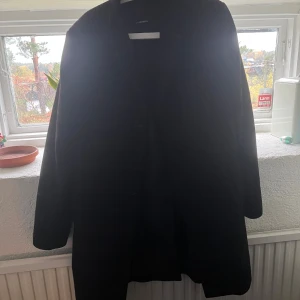 Rutig svart rock från J.Lindeberg - Snygg svart rock från J.Lindeberg med diskret röd-rutig design. Jackan har klassisk rak passform, knäppning framtill och är tillverkad i ullblandning med GORE Windstopper-foder som skyddar mot vind. Perfekt för kyliga dagar när du vill se stilren ut.