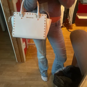 Vit handväska från Michael Kors - Snygg vit handväska från Michael Kors med guldfärgade nitar runt kanterna och matchande detaljer. Väskan har både handtag och axelrem, samt praktiska fack inuti. Stilren och boxig form, perfekt för dig som gillar lyxiga accessoarer med attityd. Nypris 2300kr!