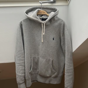 Grå hoodie från Polo Ralph Lauren - Snygg grå hoodie från Polo Ralph Lauren med klassisk broderad logga på bröstet. Tröjan har huva med snörning, stor magficka och ribbade muddar. Perfekt för kalla höst dagar och enkelt och matcha. Hör av er med minsta funderingar 