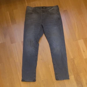 J & J Jeans - Snygga grå jeans från Jack and Jones  Slim Glenn i slim fit-modell. Jeansen har klassisk femficksdesign, raka ben och diskreta slitningar för en cool look. Perfekta för dig som gillar en smalare passform och stilren vibe.