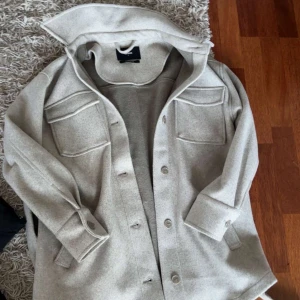 Beige overshirt med fickor - Stilren beige overshirt med stora bröstfickor och knappar framtill. Skjortan har klassisk krage, långa ärmar med manschett och är tillverkad i ett mjukt material som ger en avslappnad look.