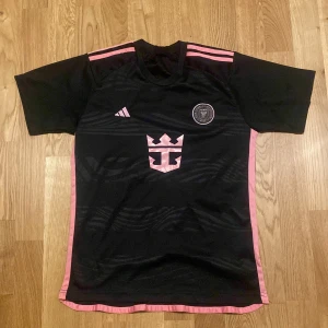 Inter Miami Messi #10 fotbollströja - Snygg svart Inter Miami fotbollströja från Adidas med rosa detaljer och Messi #10 på ryggen. Tröjan har korta ärmar, rosa ränder på axlarna och sidopaneler, samt klubbmärke och Adidas-logga på bröstet. Tillverkad i lätt och ventilerande material.