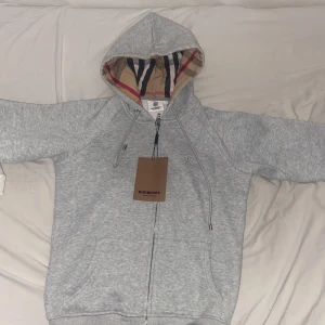 Grå hoodie från Burberry med dragkedja - Säljer en grå hoodie från Burberry med klassiskt rutigt foder i huvan och snygga snören. Tröjan har dragkedja framtill, fickor och långa ärmar. Perfekt för dig som gillar stilrena märkesplagg med en twist. Ikonisk Burberry-logga broderad på bröstet.