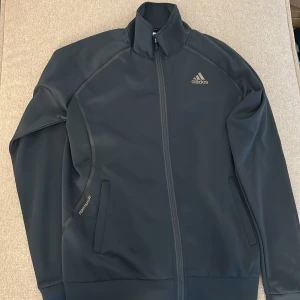  Kofta  från Adidas - Sportig kofta från Adidas med hög krage och hel dragkedja framtill. Jackan har två sidofickor och diskret Formotion 3D-detalj på ena ärmen. Tillverkad i slät polyester, perfekt för träning eller chill. Klassisk Adidas-logga på bröstet.