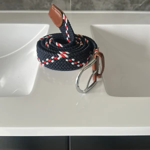 Randigt flätat bälte - Snyggt flätat bälte i marinblått med röda och vita ränder. Bältet har bruna läderdetaljer vid spännet och änden samt ett silvrigt metallspänne. Perfekt accessoar för att lyfta din outfit med lite färg och stil.