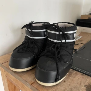 Svarta Moon Boot  - Säljer ett par klassiska svarta Moon Boot The Original med vit logga och snörning framtill. Lite sönder på en hälen men annars i gott skick! Storlek 42-44