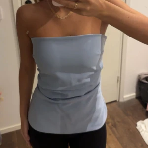 Ljusblå axelbandslös tubtopp - Säljer en stilren ljusblå tubtopp i bandeau-modell. Toppen är axelbandslös och har en slät, enkel design som sitter tight runt överkroppen. Materialet känns mjukt och har en lätt glans, perfekt för att skapa en clean look. Passar dig som gillar minimalistisk stil.