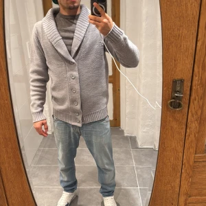 Ljusgrå kofta från Fresh Brand - Säljer en ljusgrå stickad kofta från Fresh Brand i storlek S. Koftan har bred krage, ribbade muddar och knappar framtill. Perfekt för dig som gillar chill och stilren look. Materialet är mjukt och håller värmen bra.