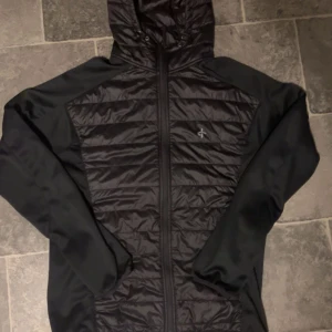 Svart vindjacka med huva - Svart vindjacka från Peak Performance med quiltad framsida och huva. Jackan har dragkedja hela vägen och ett stilrent, sportigt utseende. Perfekt för dig som gillar en clean look och vill ha något lätt att slänga på sig när det blåser.