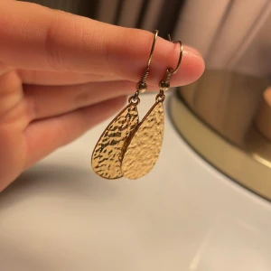 Guldiga droppformade örhängen - Snygga örhängen i droppform med hamrad yta som ger en cool textur. De är guldiga och har krokar för enkel användning. Perfekta för dig som gillar statement-smycken med lite edge och glans. Passar till alla tillfällen när du vill sticka ut lite extra.