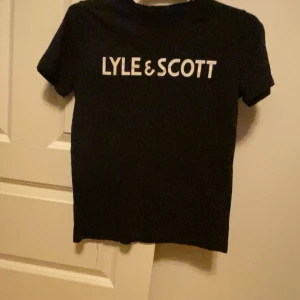 Svart t-shirt Lyle & Scott Junior - Svart t-shirt från Lyle & Scott Junior med vit logotyp tryckt på bröstet. Klassisk rund hals och korta ärmar. T-shirten har en clean och stilren look som funkar till jeans eller shorts. Materialet känns mjukt och bekvämt mot huden.