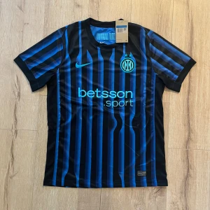 Inter Milan randig fotbollströja Nike - Snygg Inter Milan fotbollströja från Nike med klassiska svarta och blå vertikala ränder. Tröjan har korta ärmar, rund hals och tryckt sponsorlogga på bröstet. Materialet är lätt och andas, perfekt för match eller träning. Inter-emblem och Nike Swoosh i blått framtill.