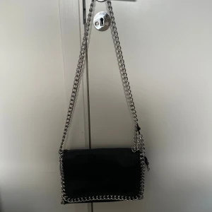 Svart väska  - Cool svart axelväska i skinn med grova silverfärgade kedjor som axelrem och runt kanterna. Tygsnöre på kedjan går lätt att klippa bort