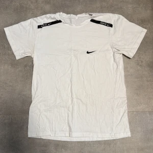 Vit Nike t-shirt med logga - Snygg vit t-shirt från Nike med svart logga på bröstet och coola svarta detaljer med Nike-tryck på axlarna. Klassisk rund hals och korta ärmar. Perfekt för dig som gillar sportig och enkel stil. Materialet är mjuk bomull som känns skönt mot huden.