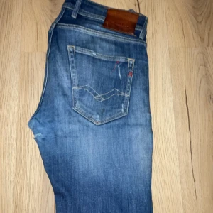 Replay Anbass jeans blå slim fit - Snygga blå Replay Anbass jeans med slim fit och slitna detaljer på benen. Klassisk femficksmodell med knappgylf och läderpatch bak i midjan. Jeansen har en cool tvättad look och är perfekta för dig som gillar streetstyle.
