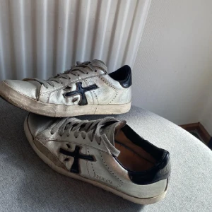Silvriga sneakers från Premiata - Säljer ett par snygga silvriga sneakers från Premiata med svarta detaljer och logga på sidan. Skorna har snörning, svart hälkappa och platt sula. Tillverkade i skinn för en lyxig känsla och passar perfekt till en streetwear-look.