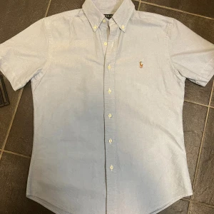 Ljusblå kortärmad skjorta Polo Ralph Lauren - Kortärmad ljusblå skjorta från Polo Ralph Lauren med klassisk button-down krage och broderad logga på bröstet. Skjortan har custom fit och stängs med vita knappar framtill. Perfekt för en clean och stilren look.