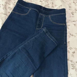 Blå denim leggings från H&M - Snygga blå denim leggings från H&M i storlek 134. Byxorna har klassiska jeanssömmar, elastisk midja och bakficka. Materialet är en mix av bomull, polyester och elastan för en bekväm och stretchig passform.