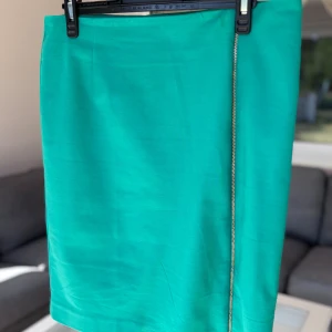  kjol med dragkedja  - Snygg turkos kortkjol från Lindex med rak passform och en cool guldfärgad dragkedja längs sidan. Kjolen är i ett slätt tyg och har en enkel, stilren look som passar perfekt till sneakers eller boots.