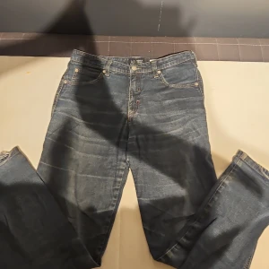 Blå Armani Jeans, storlek 30 - Snygga blå jeans från Armani Jeans med klassisk femficksdesign och logga på bakfickan. Jeansen har rak passform och är tillverkade i bomull med lite elastan för extra komfort. Perfekt för dig som gillar stilrena och tidlösa jeans. Dem har en skade på höger häl.