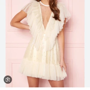 Vit kortklänning med volang och pärlor - Supersöt vit kortklänning från Ida Sjöstedt med djup v-ringning, volangdetaljer och pärlor på tyget. Klänningen har transparent mesh upptill och plisserade volanger längs axlar och fåll. Perfekt för dig som vill sticka ut på studenten! 