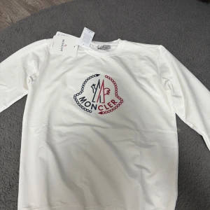 Vit sweatshirt med brodyr - Säljer en stilren vit sweatshirt  i bomull med broderad logga i rött och marinblått framtill. Tröjan har rund halsringning och långa ärmar, perfekt för dig som gillar exklusiva och cleana plagg.