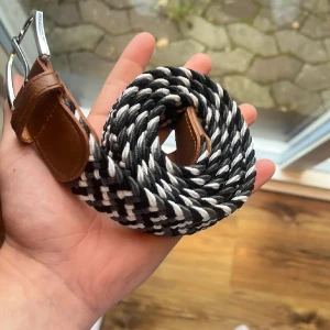 Svartvitt flätat bälte med skinn - Säljer ett snyggt flätat bälte i svart och vitt med bruna skinn-detaljer vid spännet. Bältet har ett silvrigt metallspänne och är perfekt för att ge outfiten lite edge. Passar både jeans och chinos.