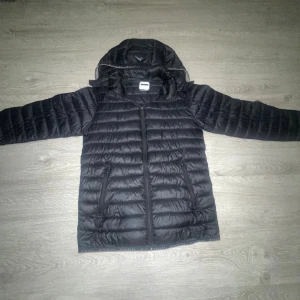 Svart dunjacka från McKinley med huva - Säljer en svart dunjacka från McKinley med huva och dragkedja framtill. Jackan är quiltad med två fickor med dragkedja och har en stilren look. Perfekt för kyliga dagar och enkel att matcha med allt.