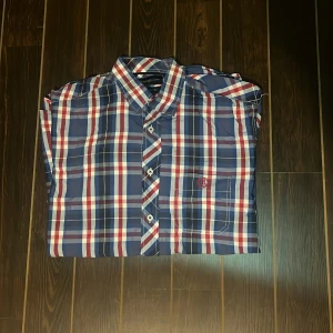 Rutig skjorta från Henri Lloyd M - Snygg rutig skjorta från Henri Lloyd i klassisk passform. Skjortan har blå, röda och vita rutor, bröstficka med broderad logga och coola knappar med brittisk flagga. Perfekt för dig som gillar stilren och tidlös design.