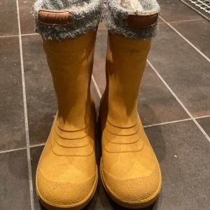 Kavat Gula fodrade stövlar med grov sula - Säljer ett par gula stövlar med grov sula och grått, mjukt foder upptill. Stövlarna har en rund tå och är tillverkade i syntetmaterial med detaljer i skinn vid kanten. Perfekta för regniga dagar och höstväder.