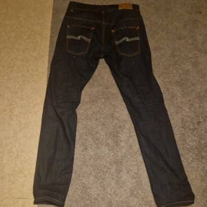Mörkblåa Nudie Jeans - Säljer ett par mörkblå jeans från Nudie Jeans med klassisk orange söm och snygg vit brodyr på bakfickorna. Jeansen har rak passform och är tillverkade i kraftig denim av bomull. Perfekta för dig som gillar stilrena och tidlösa jeans. W31 L32