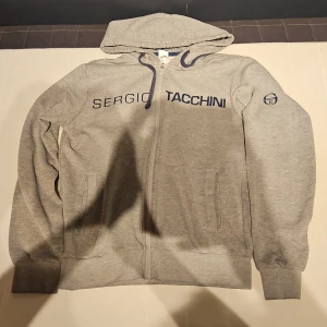 Grå hoodie från Sergio Tacchini S - Grå hoodie från Sergio Tacchini i storlek S. Tröjan har dragkedja framtill, huva med snörning och två fickor. Stort tryck med logga över bröstet och emblem på ärmen. Mjuk insida och klassisk passform, perfekt för chill dagar. Perfekt som huligan/ultras tjocktröja.