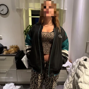 Oversized svart och grön bomberjacka - Säljer en oversized skinnjacka i svart och grön med vita detaljer och stora 'Los Angeles' tryckt på ryggen. Jackan har dragkedja framtill, långa ärmar och sportig collegekänsla. Perfekt statement-plagg för dig som gillar streetstyle.