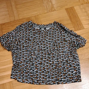 Nike Just Do It kort t-shirt - Nike t-shirt i svart med all-over print av 'Just Do It.' i vitt och små orangea Nike-loggor. Kortärmad och croppad modell, perfekt för en sportig och trendig look. Mjuk och skön bomullskänsla, passar dig som gillar streetwear.